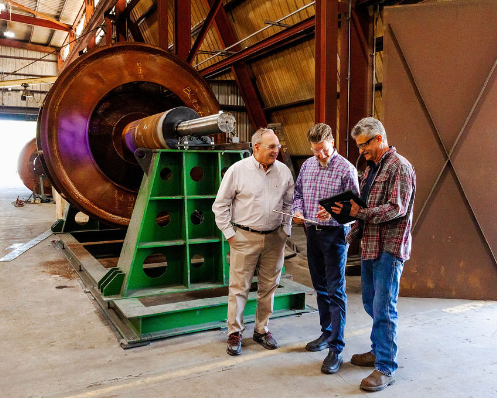 three men looking at a tablet and clipboard in front of a fan